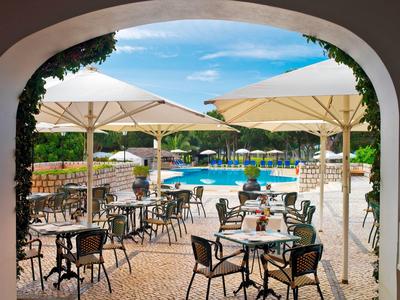 Sonnige Poolterrasse mit Tischen, Stühlen und großen Sonnenschirmen in Hotelanlage.