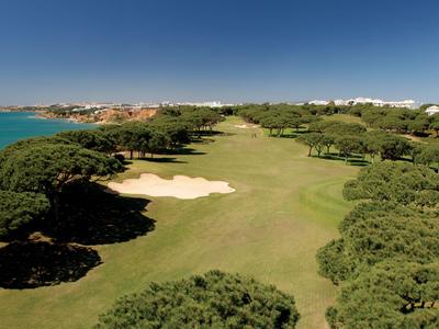 Vista de un campo de golf costero con bunkers de arena y árboles bajo un cielo azul claro.