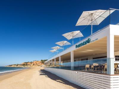 Modernes Hotel mit Terrasse und Sonnenschirmen direkt am Sandstrand unter klarem blauem Himmel.