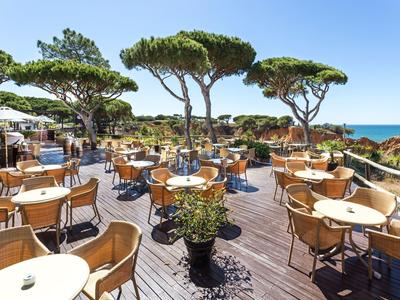 Offene Terrasse mit Tischen und Stühlen unter Kiefern und klarem Himmel am Meer.