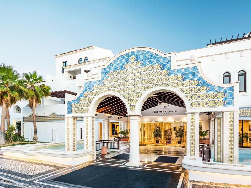 Eingang eines weißen Hotels mit blauen Details und Palmen unter klarem Himmel.