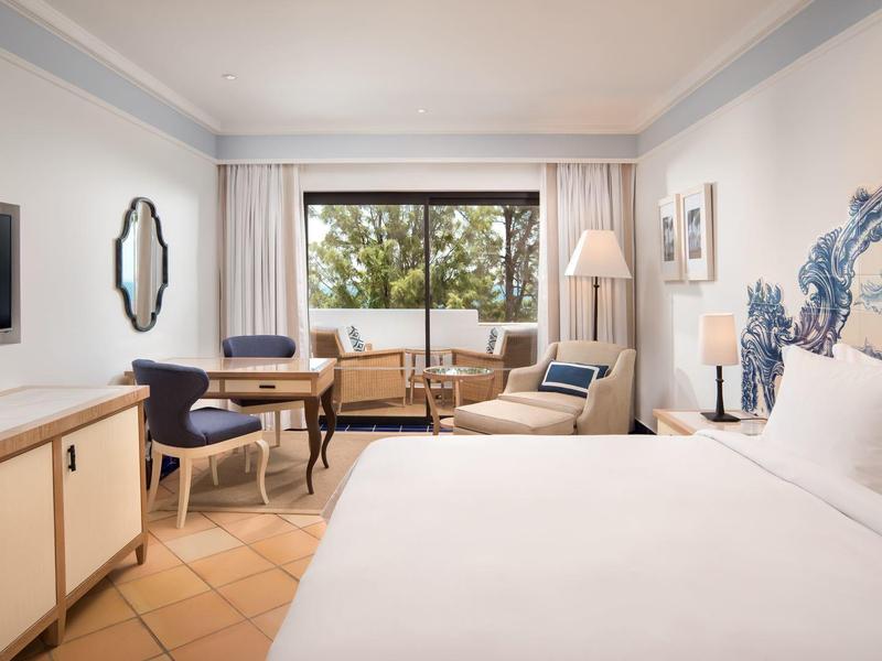 Helles Hotelzimmer mit großem Bett, Schreibtisch, Stühlen und Blick auf grüne Bäume.