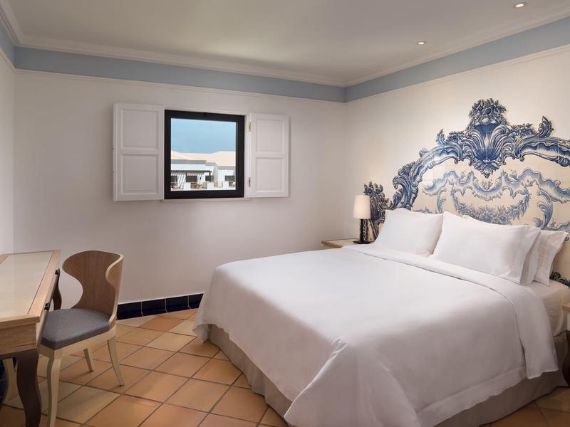 Helles Hotelzimmer mit großem Bett, dekorativem Kopfteil, Schreibtisch und Fenster mit Stadtblick.