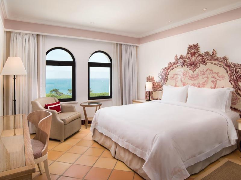 Helles Gästezimmer mit Boxspringbett, Vintage-Kopfteil, zwei Stühlen, Schreibtisch und Meerblick.