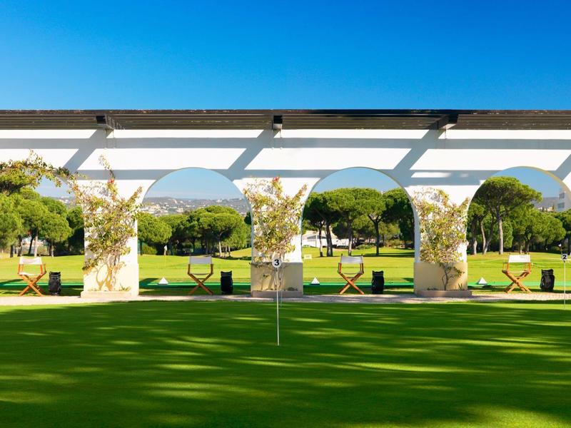 Grüner Golfplatz mit weißer Pergola und Bäumen im Hintergrund bei sonnigem Wetter.