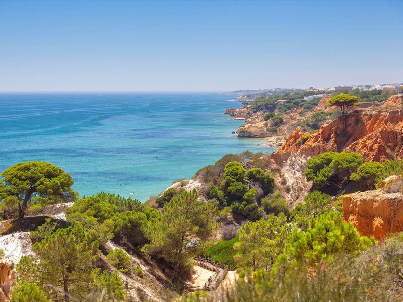 Küste mit roten Felsen, grüner Vegetation und blauem Meer unter klarem Himmel.