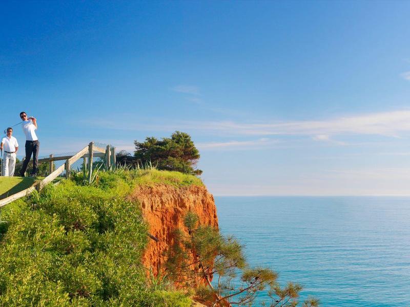 Zwei Personen stehen auf einer grünen Klippe mit Blick auf das blaue Meer unter klarem Himmel.