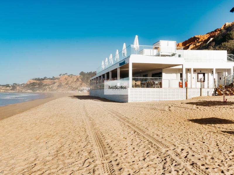 Modernes weißes Gebäude am Sandstrand mit Bergen im Hintergrund unter klarem Himmel.
