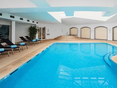 Hallenbad mit hellem blauen Wasser, Holzfußboden, Liegen und großen Fenstern in Weiß.