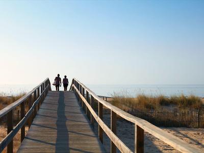 Zwei Personen gehen auf einem hölzernen Steg zum Strand bei klarem Himmel und ruhigem Meer.