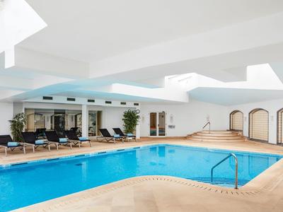 Innenpool mit blauem Wasser, Liegestühlen, Pflanzen und großen Fenstern in hellem Raum.