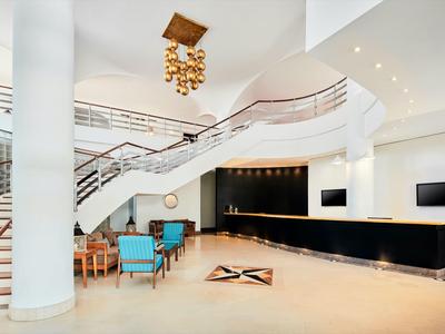 Helle Hotellobby mit geschwungener Treppe, eleganter Deckenleuchte und Sitzbereich mit blauen Stühlen.