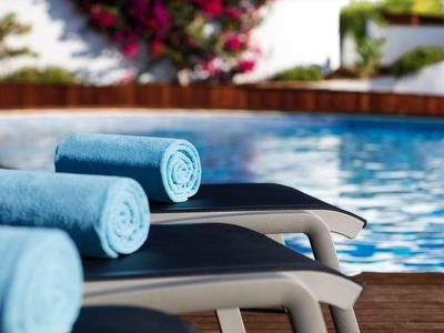 Drei blau gerollte Handtücher liegen auf schwarzen Poolliegen neben klarem Schwimmbecken.