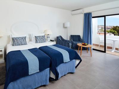 Helles Hotelzimmer mit zwei Betten, blau-weißer Bettwäsche, Sitzbereich und Balkon mit Außenblick.