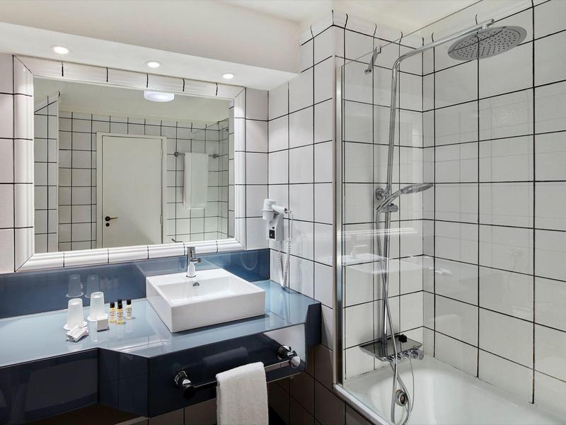 Helles Badezimmer mit weiß gekachelten Wänden, großer Spiegel, Waschtisch und Dusche mit Glasabtrennung.