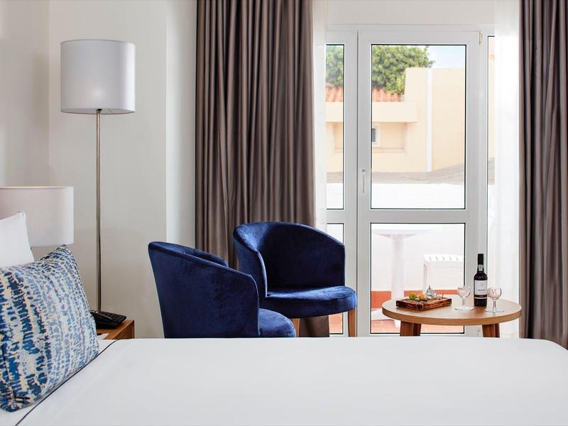 Helles Schlafzimmer mit zwei dunkelblauen Sesseln, weißem Bett und Tisch mit Wein und Obst.