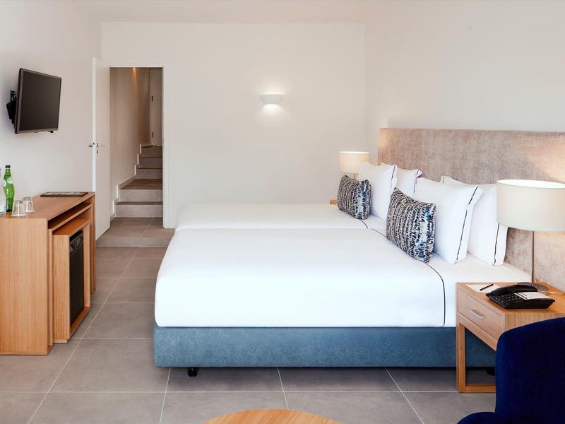 Helles Hotelzimmer mit zwei Doppelbetten, TV an der Wand und modernem Holzmöbel-Set.