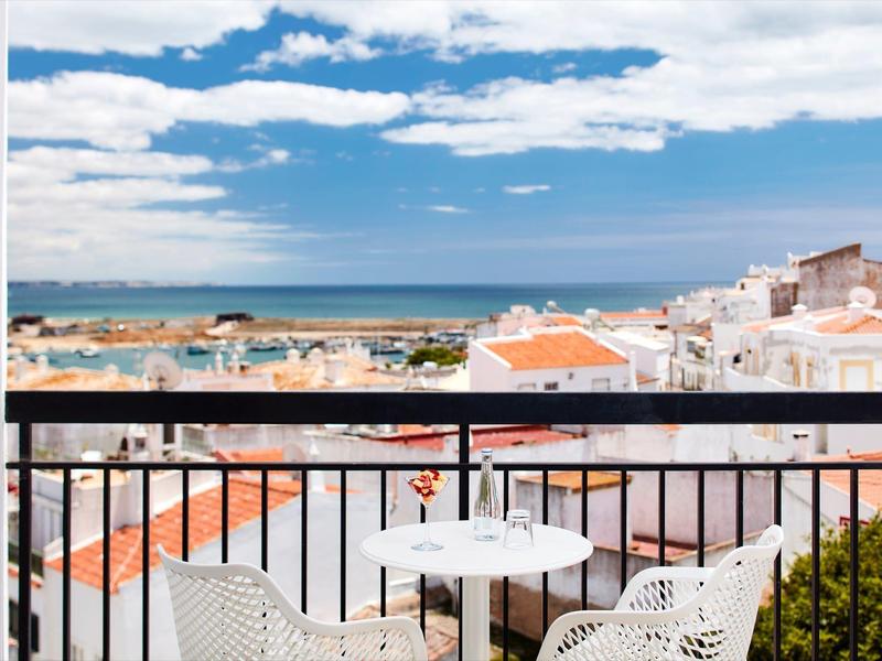 Balkon mit weißem Tisch und Stühlen, Blick auf rote Dächer und blaues Meer unter Wolkenhimmel.