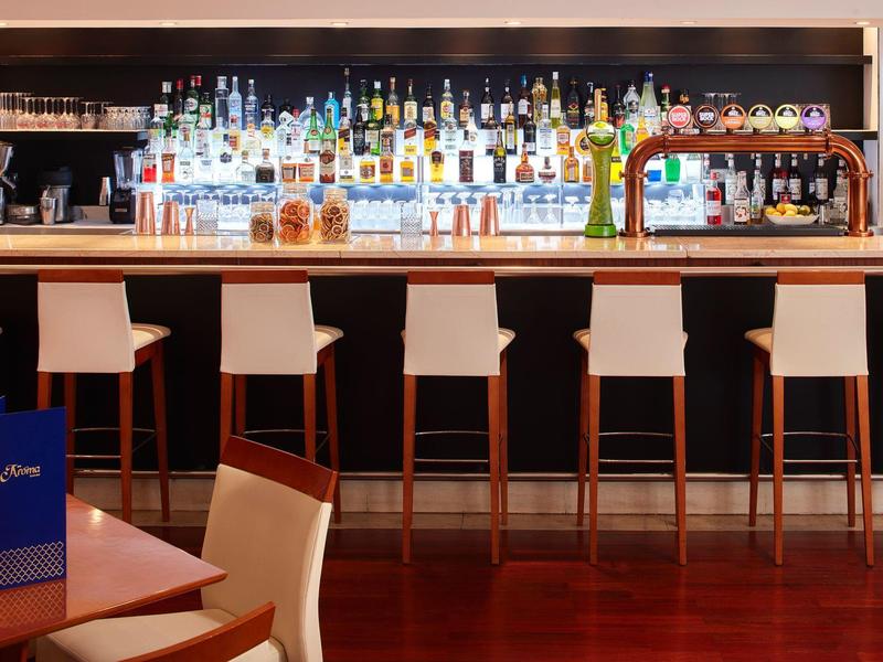 Moderne Bar mit hohen weißen Stühlen, Holztheke und vielen bunten Spirituosenflaschen im Regal.