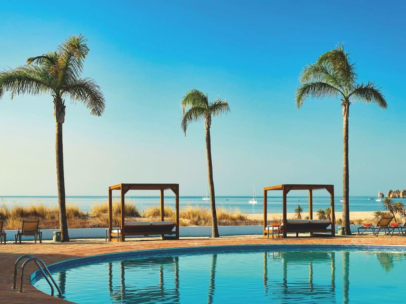 Ein klarer Pool vor dem Strand, umgeben von Palmen und Sonnenliegen bei blauem Himmel.