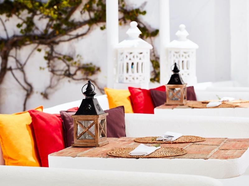 Weiße Tische mit braunen Platzsets, bunt gefüllte Kissen und dekorative Laternen in hellem Raum.