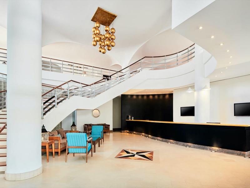 Helle Hotellobby mit geschwungener Treppe, eleganter Deckenleuchte und Sitzbereich mit blauen Stühlen.