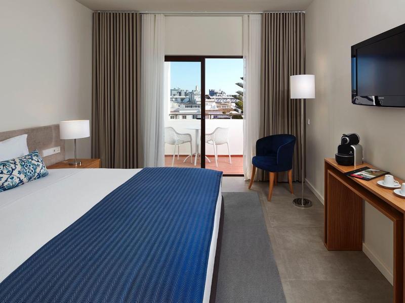 Modernes Hotelzimmer mit großem Bett, blauer Tagesdecke, Balkon und Fernseher an der Wand.