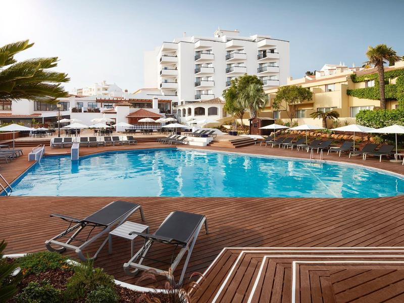 Großer Pool mit Sonnenliegen, Palmen und mehrstöckigen weißen Gebäuden im Hintergrund.