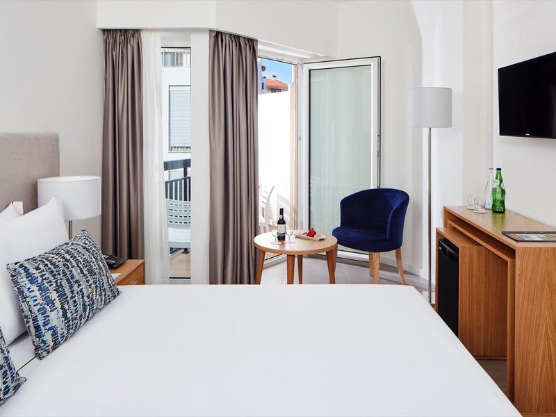 Helles, modernes Schlafzimmer mit großem Bett, Balkon, blauer Sitzgelegenheit und Wandfernseher.