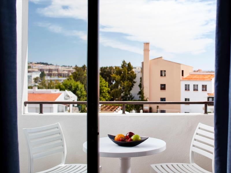 Blick durch Fenster auf Balkon mit weißem Tisch, Stühlen und Obstschale, dahinter Häuser und Himmel.