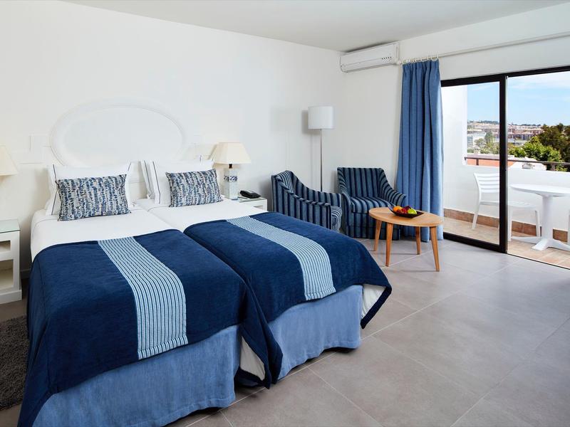 Helles Hotelzimmer mit zwei Betten, blau-weißer Bettwäsche, Sitzbereich und Balkon mit Außenblick.