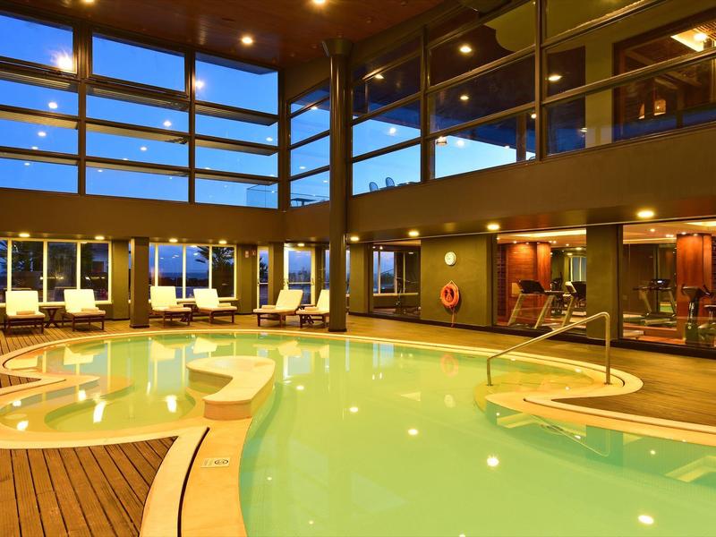 Moderner Innenpool mit Holzverkleidung, bequemen Liegestühlen und großes Fensterfront bei Abendlicht.