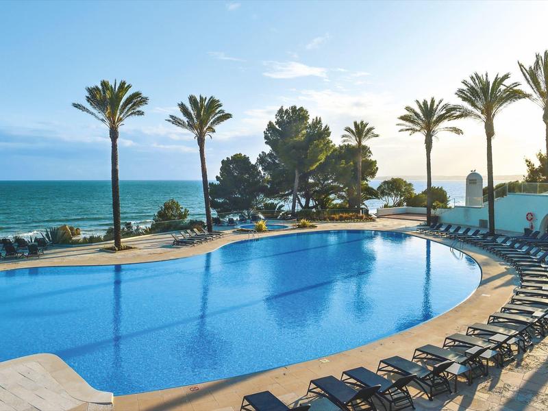 Großer Pool mit blauer Wasserfläche, Palmen und Sonnenliegen am Meer unter blauem Himmel.