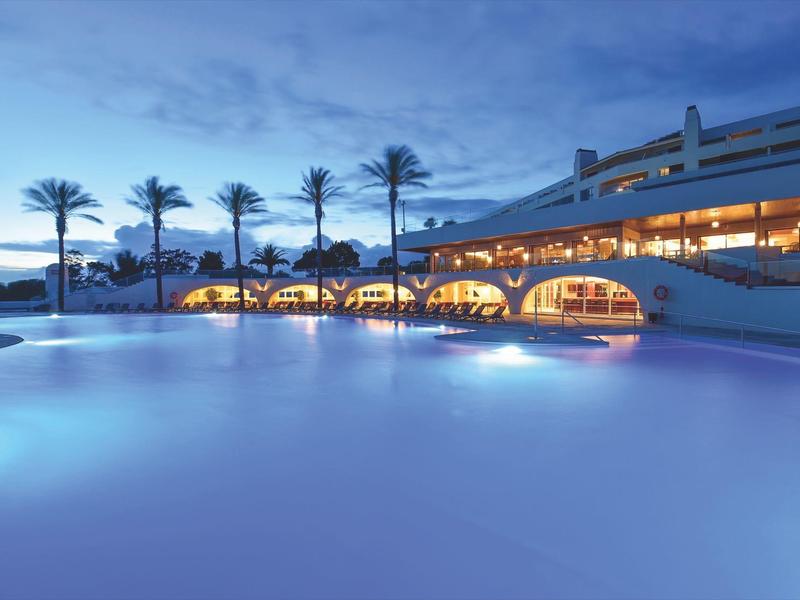 Großer Außenpool bei Abenddämmerung, Palmen und beleuchtetes, rundes Gebäude im Hintergrund.