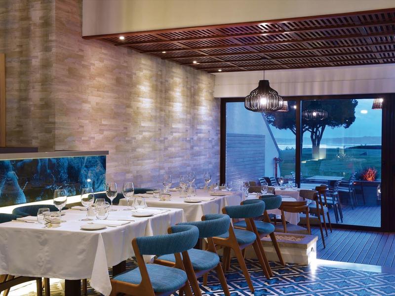 Eleganter, heller Speiseraum mit Holztischen, Stühlen und Meerblick bei Sonnenuntergang.