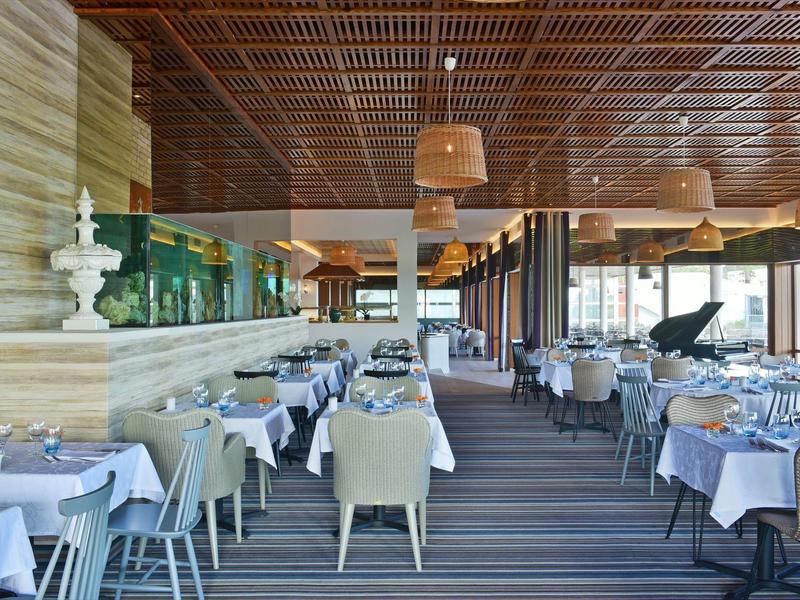 Großes, helles Restaurant mit gedeckten Tischen, Stühlen, Holzdecke und Klavier am Fensterbereich.