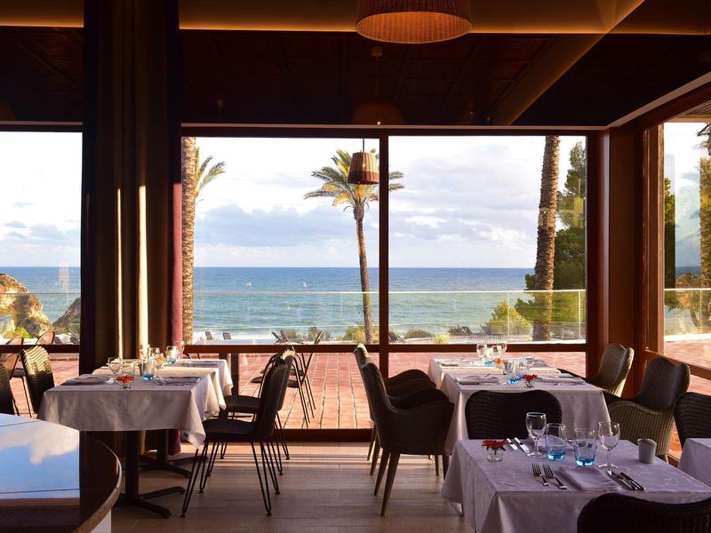 Restaurant mit gedeckten Tischen, Stühlen und Aussicht auf Meer, Palmen und blauen Himmel.