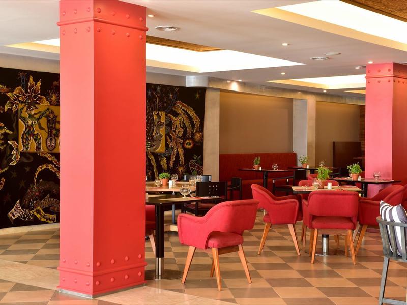 Moderner Gastraum mit roten Säulen, roten Sesseln und gedeckten Tischen auf gemusteretem Boden.