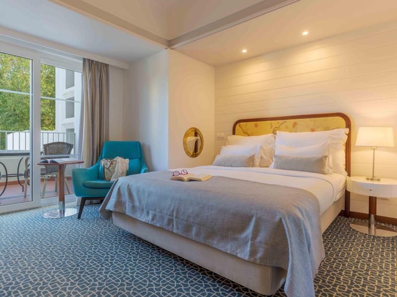 Helles Schlafzimmer mit großem Bett, blauem Sessel, Teppichboden und Balkon mit Sitzgruppe.