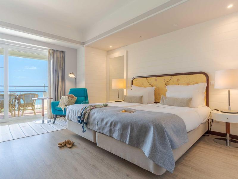 Helles Hotelzimmer mit großem Bett, blauen Sesseln, Holzböden und Balkon mit Meerblick.