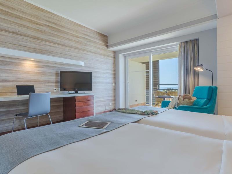 Modernes Hotelzimmer mit Doppelbett, Schreibtisch, Fernseher, blauem Sessel und Balkon mit Meerblick.