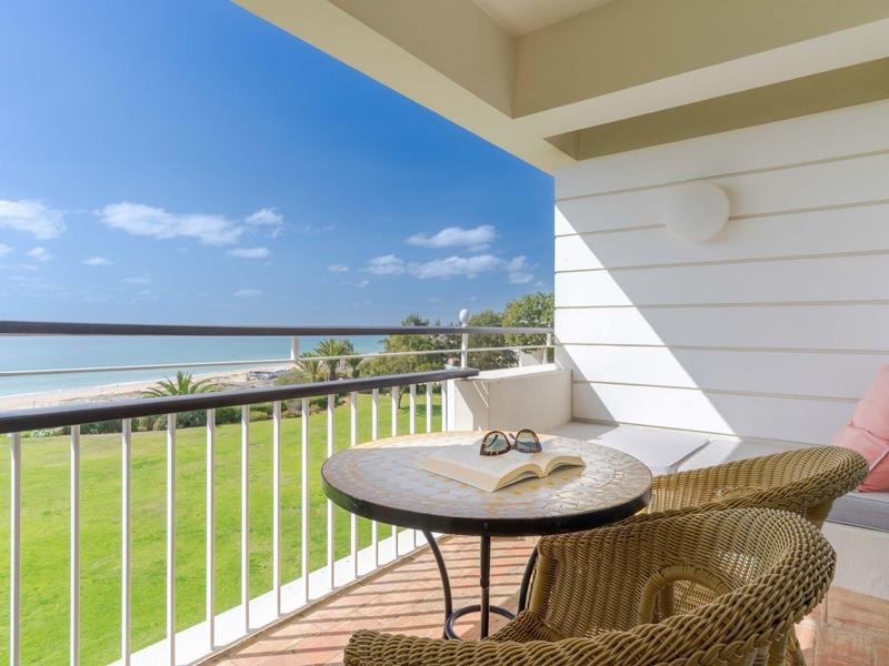 Balkon mit Tisch, zwei Stühlen, Blick auf grünen Rasen, Strand und blaues Meer unter klarem Himmel.