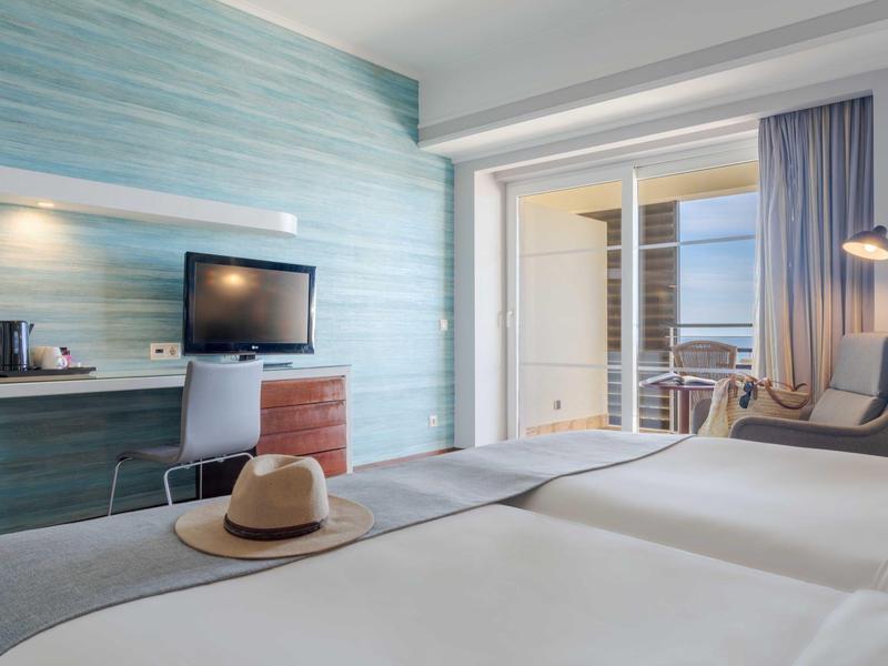 Modernes Hotelzimmer mit zwei Betten, TV, Schreibtisch, Balkon und Stuhl mit Stehlampe.