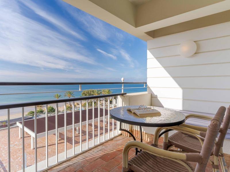 Balkon mit rundem Tisch, zwei Stühlen und Blick auf Meer, Leuchtturm und blauen Himmel.