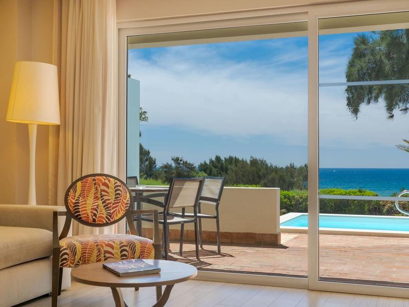 Helles Wohnzimmer mit Sesseln und Lampe, großer Glastür mit Blick auf Terrasse, Pool und Meer.