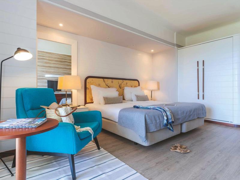 Modernes Schlafzimmer mit grauem Bett, blauem Sessel, Teppich und weißen Wänden in hellem Holz.