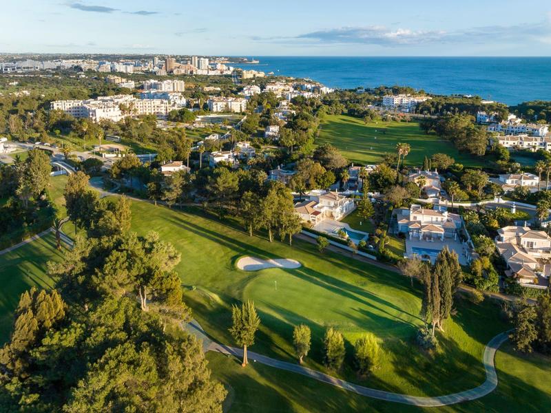 Grüner Golfplatz mit Bunkern, umgeben von Häusern, Bäumen und Blick auf das blaue Meer.