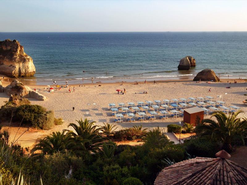 Breiter Sandstrand mit Sonnenliegen, Felsen im Wasser und Palmen im Vordergrund am Meer.