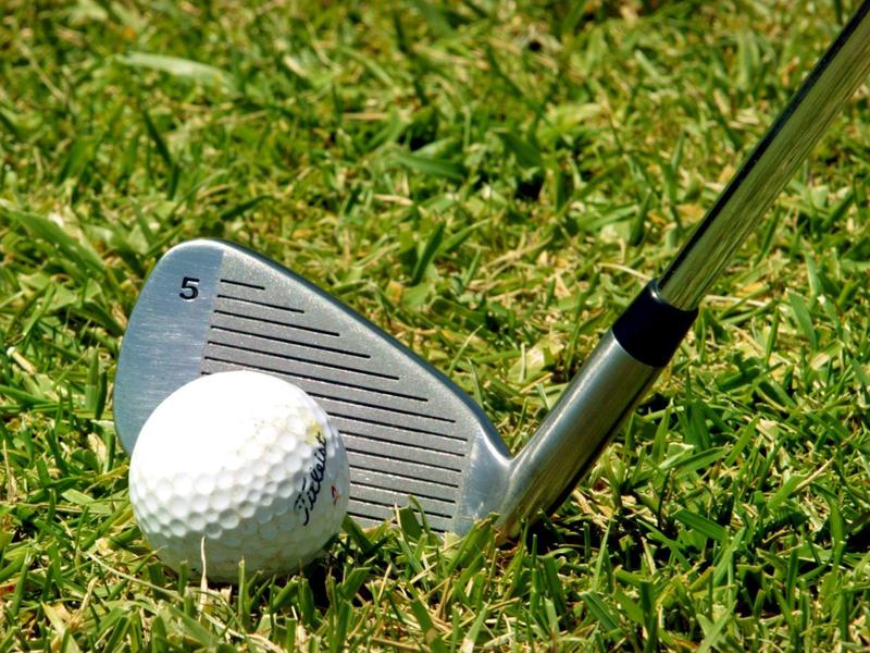 Ein Golfball liegt in kurzem grünen Gras vor einem silbernen Golfschläger mit der Zahl 5.