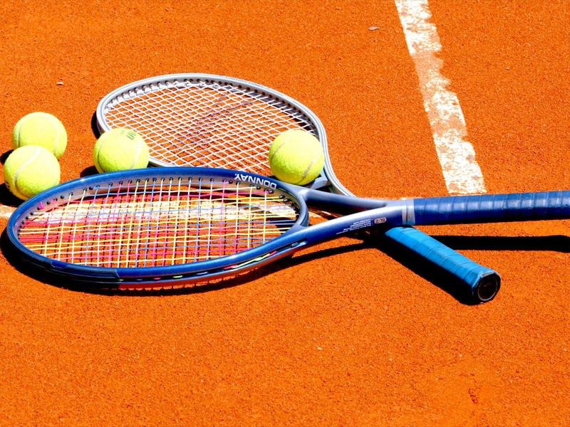 Zwei blaue Tennisschläger und fünf gelbe Tennisbälle liegen auf einem roten Sandplatz.